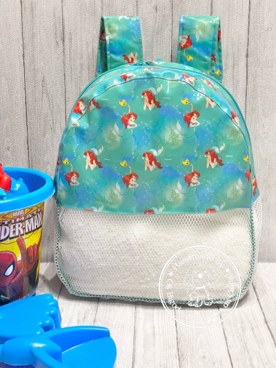Mochila infantil azul com padrão da Pequena Sereia e malha branca ao lado de balde e pá do Spider-Man