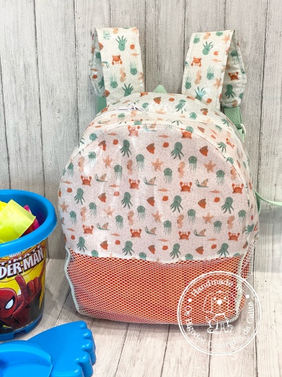 Mochila infantil colorida com padrão de animais marinhos, alças almofadadas e bolso em tela laranja ao lado de balde de praia do Homem-Aranha.