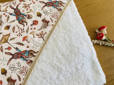 Manta para bebé com padrão de coelhos e elementos da natureza num lado e tecido branco felpo no outro lado