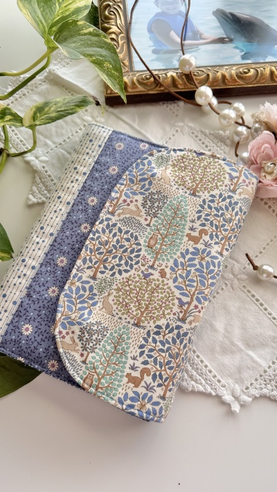 Carteira ou estojo de tecido com padrão floral e árvores junto a moldura dourada, colar de pérolas e rosa artificial sobre pano branco bordado