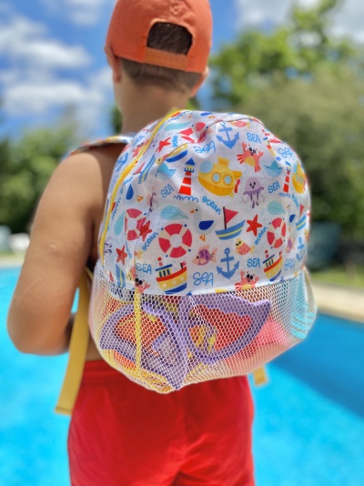 Mochila infantil colorida com padrão marinho e parte inferior em tela, vista nas costas de uma criança junto a uma piscina