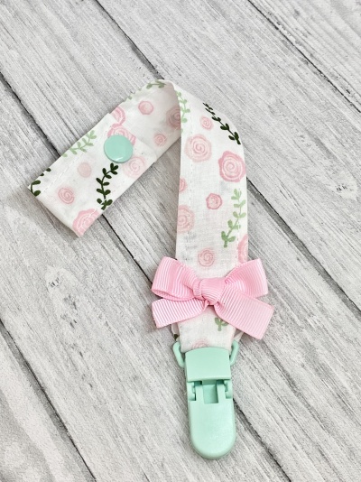 Prendedor de chupeta com padrão floral rosa e verde, botão e clip verde pastel, laço rosa, sobre fundo de madeira cinza claro