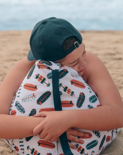 Criança com boné preto abraça mochila branca com padrão de pranchas de surf na praia.