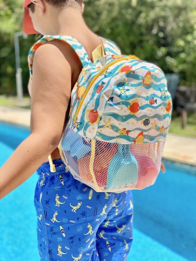 Mochila infantil colorida com padrão de peixes e rede, usada por criança junto a piscina