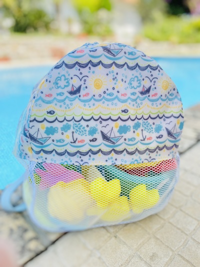 Mochila pequena estampada com padrão de barcos e ondas, contendo brinquedos de banho em bolso de rede, próxima a uma piscina
