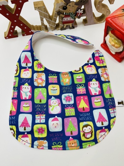 Babete azul com padrão colorido de Natal e texto em inglês, com fecho de velcro, junto a decoração natalícia em madeira e figura de Pai Natal.
