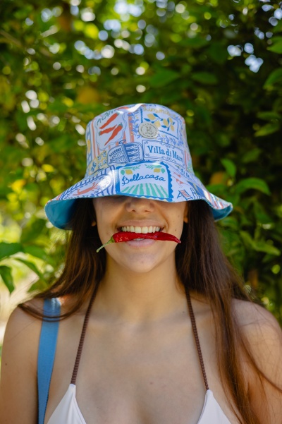 Chapéu bucket estampado colorido com palavras, pessoa com malagueta vermelha na boca e fundo de folhas verdes