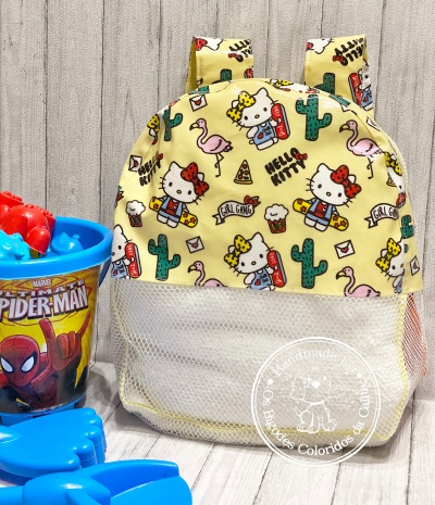 Mochila infantil amarela com padrão da Hello Kitty com cactos, flamingos e cupcake ao lado de brinquedos e copo do Spider-Man