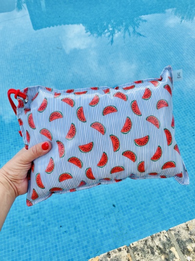 Mala pouch azul claro com padrão de fatias de melancia vermelha e verde segurada à frente de uma piscina azul.