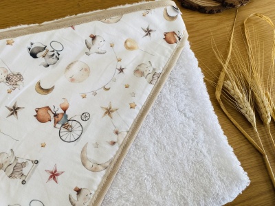 Toalha branca com capuz estampado de estrelas, luas e animais sobre madeira clara com ramos secos