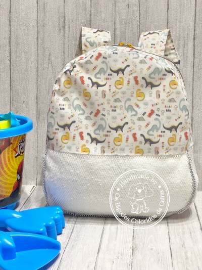 Mochila infantil estampada com dinossauros coloridos e brinquedos azuis ao lado