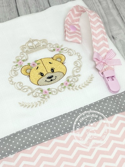 Fralda branca bordada com ursinho e faixa cinza de pontos brancos, junto com peçonha rosa com padrão zigzag e laço.