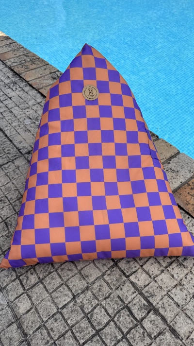 Puff triangular com padrão xadrez laranja e roxo ao lado de piscina.