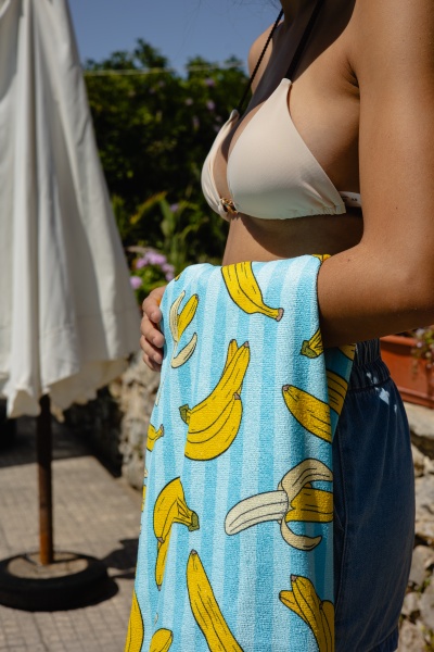 Toalha de banho azul com bananas amarelas segurada por pessoa com fato de banho bege ao ar livre