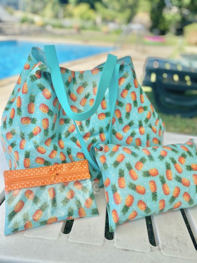 Conjunto de mochilas e bolsas com padrão de ananás em fundo azul, sobre uma mesa branca junto a piscina