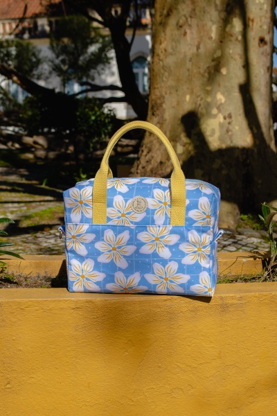 Mala de mão azul com flores brancas e amarelas, alças amarelas, etiqueta redonda, muro amarelo ao fundo