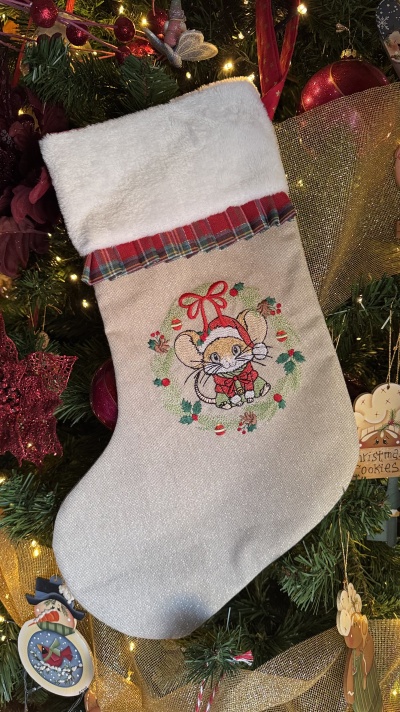 Meia de Natal bege com bordado de rato e punho branco, pendurada em árvore de Natal decorada.