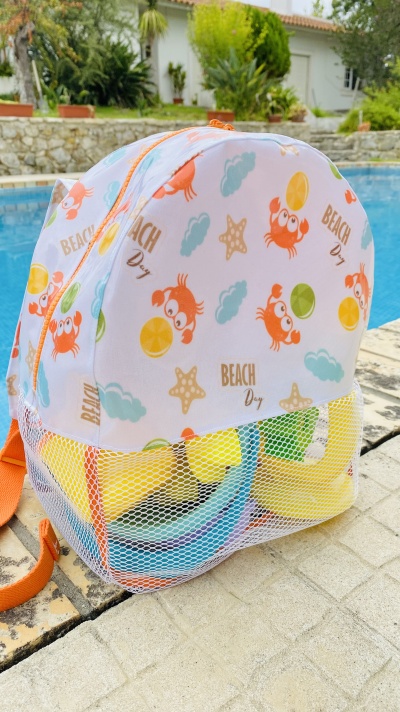 Mochila infantil branca com estampado colorido de caranguejos e estrelas-do-mar, bolso de rede com brinquedos, encostada no chão junto a piscina.