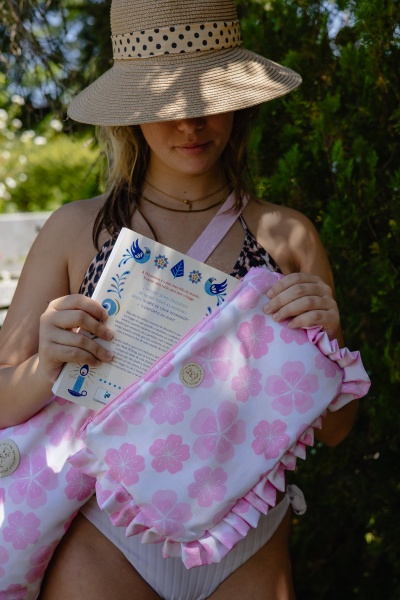 Mulher com chapéu de palha e roupa de banho segurando almofada rosa com flores e um livro.