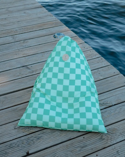Pufe triangular verde com padrão de quadrados branco e etiqueta circular de couro, posto em deque de madeira junto à água