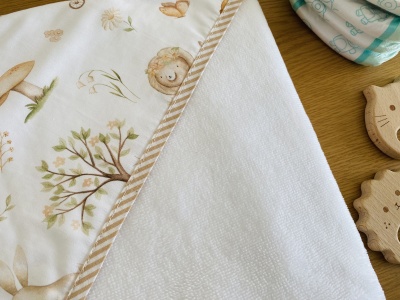 Toalha branca para bebé com padrão decorativo e borda às riscas sobre superfície de madeira clara com brinquedos de madeira