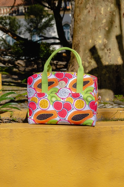 Mala de mão com padrão de frutas tropicais e alças verdes sobre muro amarelo