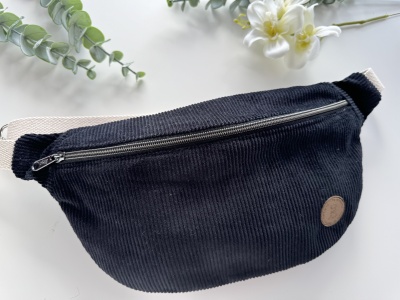 Bolsa tipo cinto preta de veludo cotelê com zip e alça bege sobre mesa branca com plantas