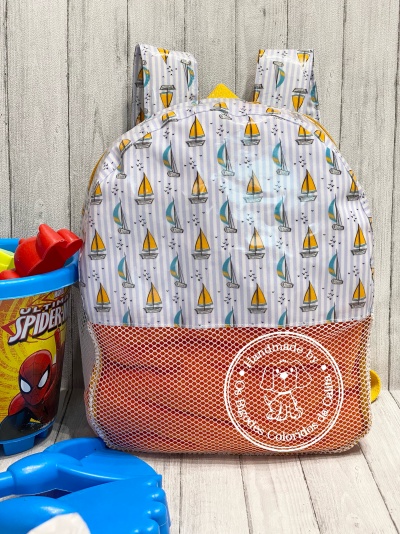 Mochila branca com veleiros amarelos e azuis e bolso laranja em malha, ao lado de balde do Homem-Aranha com brinquedos