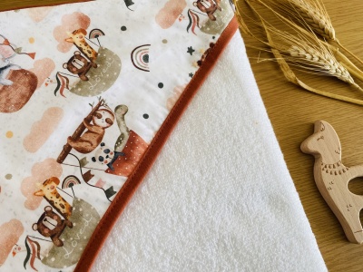 Fralda de bebé branca com borda laranja e estampado de animais, em mesa de madeira com espigas de cereal e brinquedo de madeira