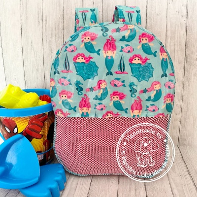 Mochila infantil azul com estampa de sereias e bolso rosa, junto a brinquedos de praia coloridos no chão de madeira clara