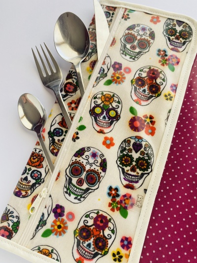 Estojo de tecido branco com caveiras coloridas e flores com talheres de metal e fundo rosa com bolinhas brancas