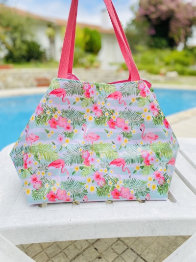 Saco colorido com padrão de flamingos, flores e folhas sobre mesa branca ao lado de piscina