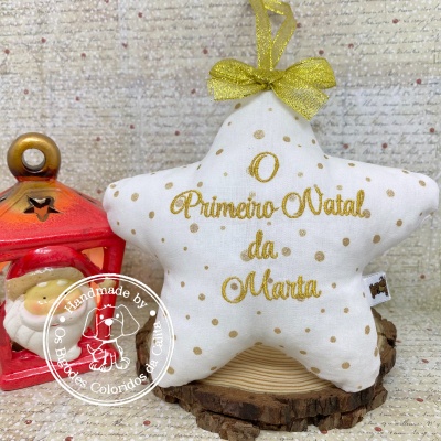 Ornamento de estrela branco com pontos dourados e texto dourado sobre suporte de madeira, ao lado de lanterna vermelha com boneco de Papai Noel