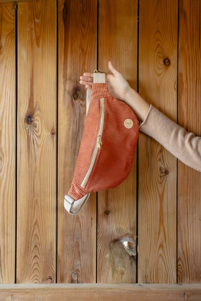 Mochila terracota em tecido canelado com fecho dourado e alças largas pretas e cinzentas segurada por uma mão