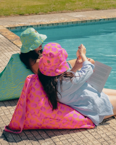 Dupla feminina sentada em puffs coloridos junto à piscina, chapéus combinando e roupa casual.