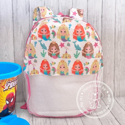 Mochila infantil com padrão de sereias e rede branca ao lado de copo azul do Homem-Aranha