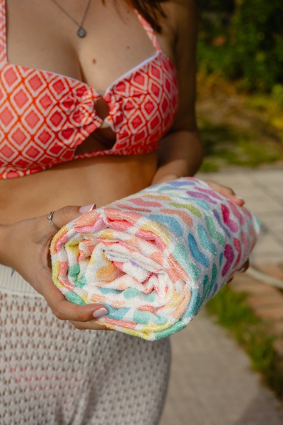 Toalha colorida enrolada com padrão de ondas, segurada por pessoa com biquíni vermelho e saia branca em ambiente exterior