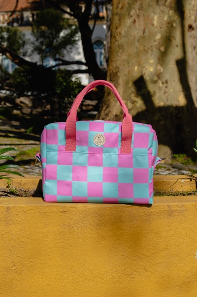 Mala de tecido azul e rosa com padrão xadrez e alças rosa sobre muro amarelo ao ar livre