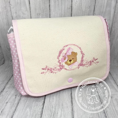 Bolsa organizadora bege com bordado de urso e detalhes cor-de-rosa e branco sobre fundo de madeira clara