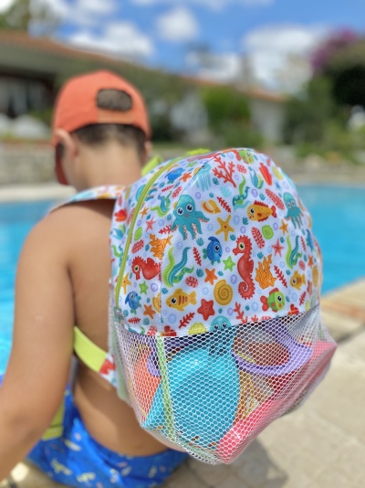 mochila infantil colorida com tema marinho e bolso de rede com brinquedos de praia usada por criança junto à piscina