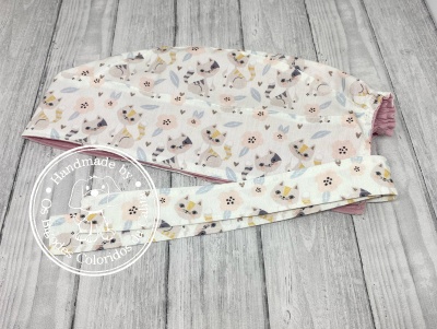Touca cirúrgica branca com padrão de gatos e flores, com faixa e interior rosa em fundo de madeira cinza
