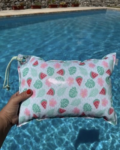 Mala laminada com padrão de melancias, folhas e flores, segurada sobre piscina azul.