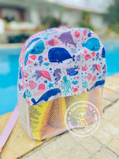 mochila infantil com padrão de animais marinhos coloridos e parte inferior em rede branca, junto a piscina