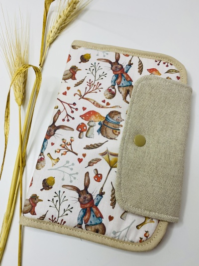 Capa de tecido com padrão de coelhos, cogumelos e plantas, com aba bege e fecho de botão, ao lado de caules de trigo.