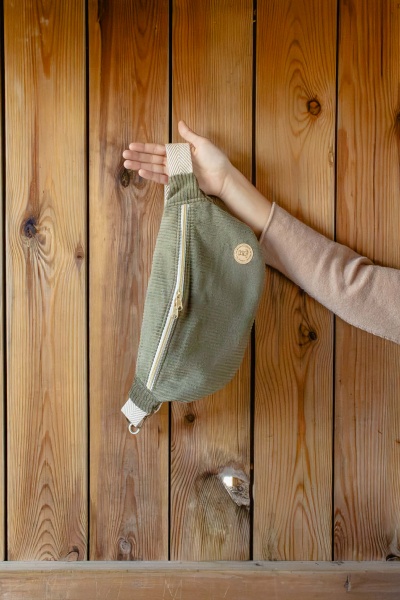 Mochila verde tipo pochete com fecho bege e alça bege, suspendida por uma mão com manga rosa claro em fundo de madeira