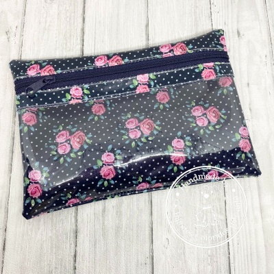Estojo transparente com padrão de rosas cor-de-rosa e bolinhas brancas sobre fundo azul escuro, com fecho de correr azul escuro.