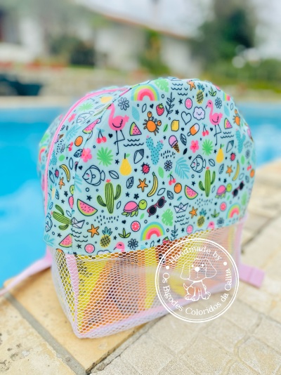 Mochila pequena colorida com padrão de flamingos, frutas e cactos, parte inferior em rede branca, junto a piscina.