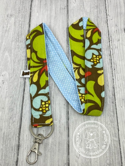 Lanyard colorido com padrão floral e bolinhas, com gancho metálico, sobre superfície de madeira clara.