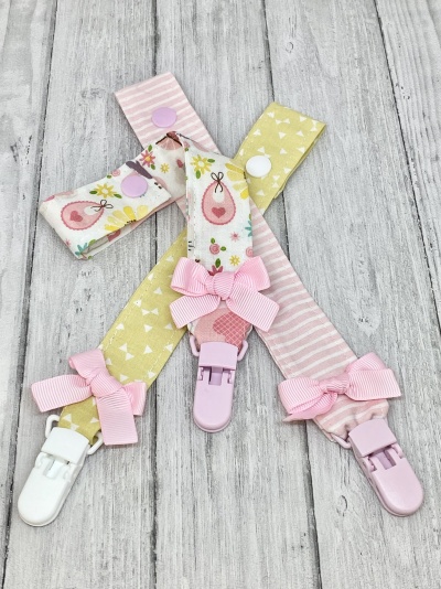 Três fitas para chupeta com padrões coloridos, presilhas plásticas e laços cor-de-rosa, sobre fundo cinzento de madeira.