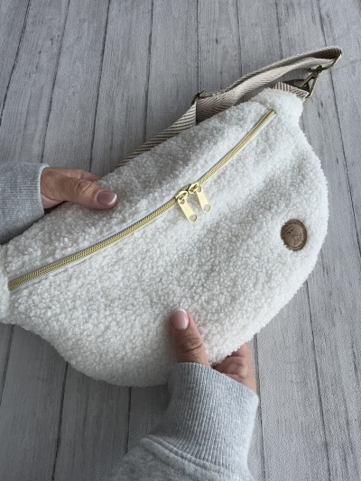 Bolsa felpuda branca com fecho dourado e alça às riscas, segurada sobre superfície cinzenta